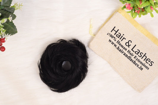 Black messy bun hair extension on white surface with Hair & Lashes pouch and decorative flowers